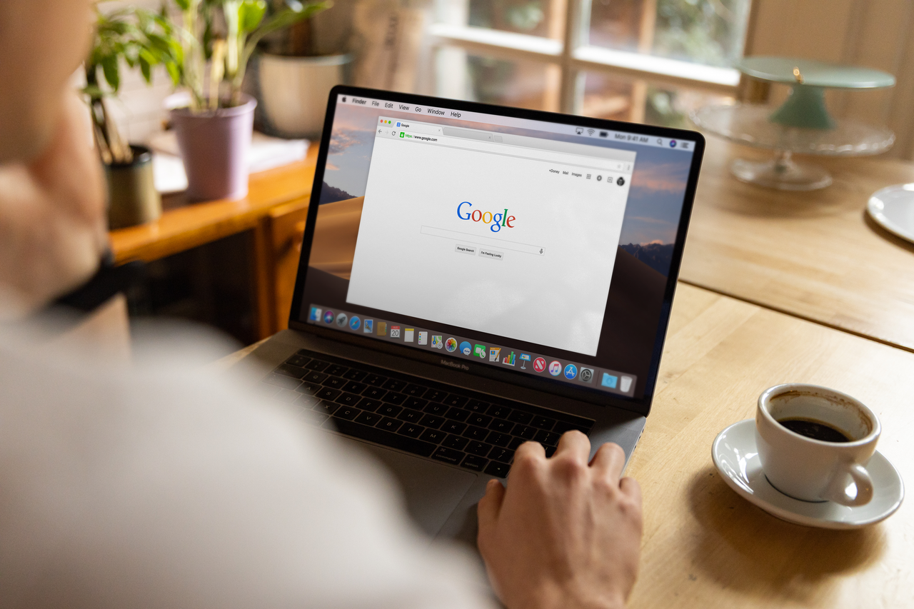 Un adulte utilise un ordinateur portable à l’intérieur, en naviguant sur Google, assis à une table en bois avec un café.